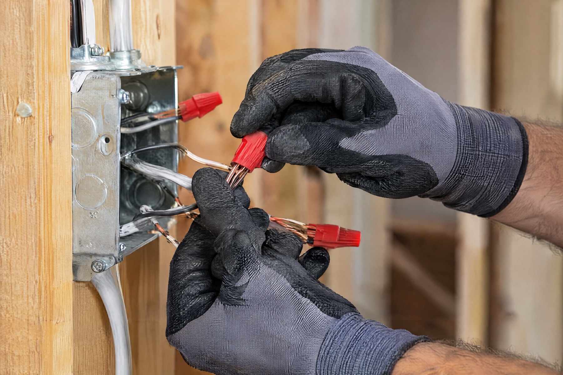 Gloved hands twisting red wire nuts onto wires at a metal junction box fixed to a wooden stud