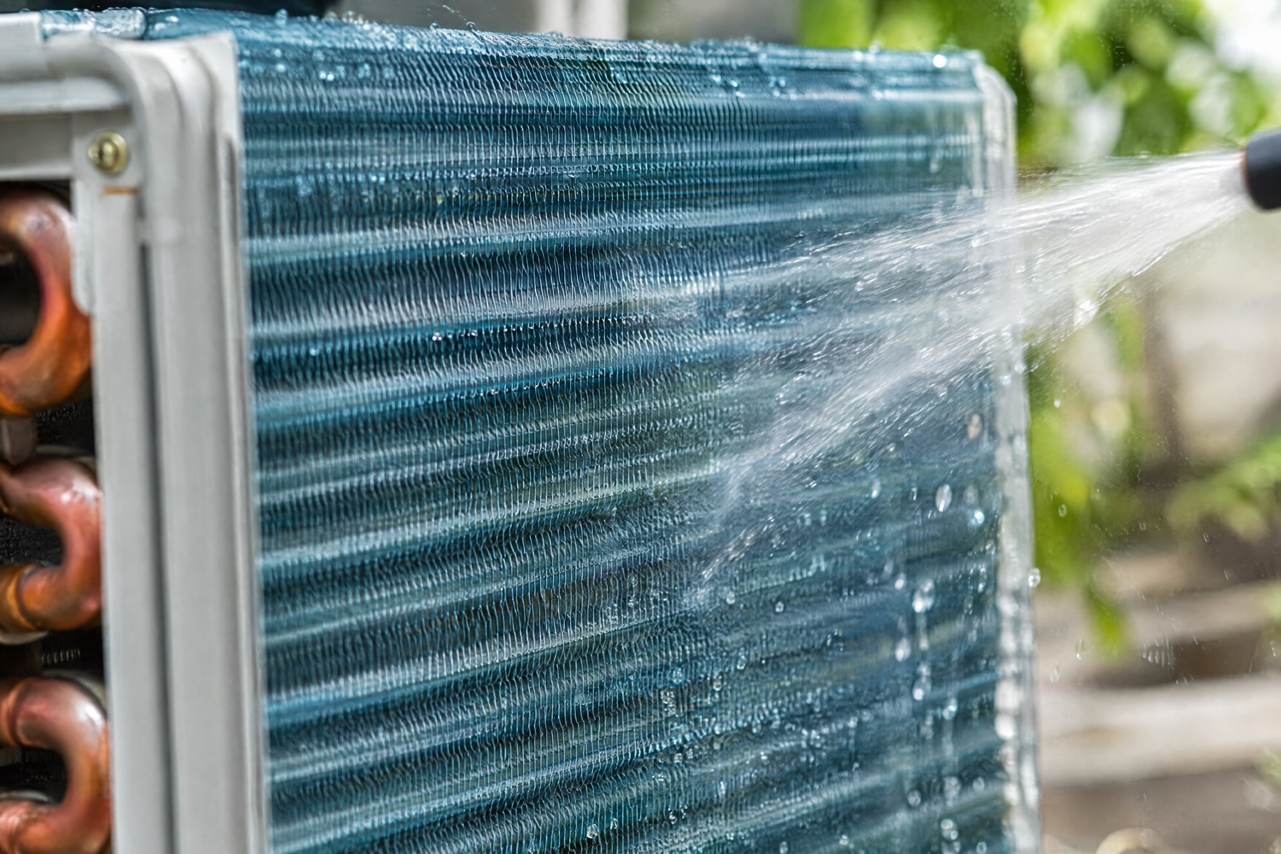 Water from a hose rinsing the blue aluminium fins of an outdoor condenser coil, with copper tubing visible at the edge