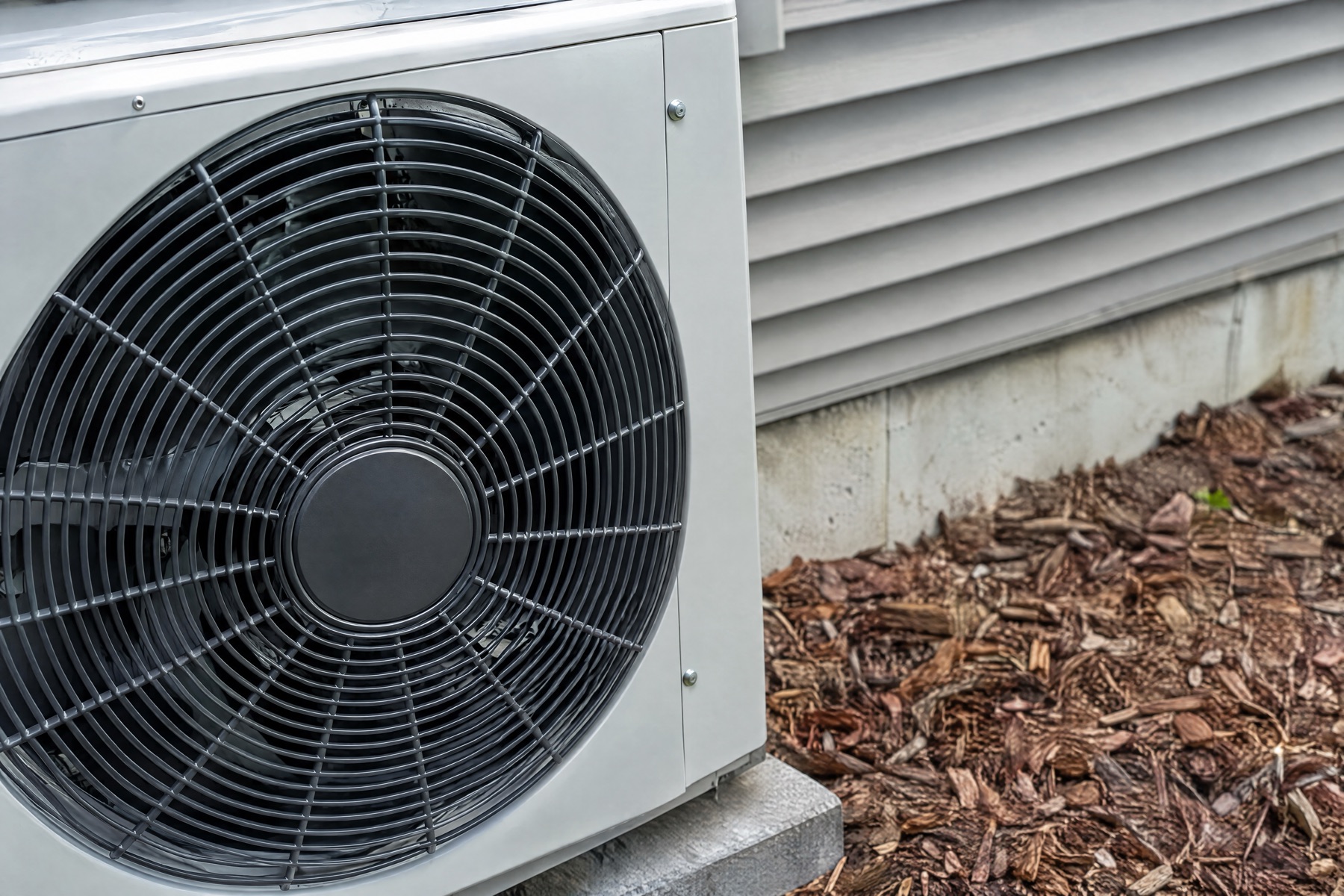 Close-up of a heat pump's outdoor unit with its round black fan grille, set against grey siding and bark mulch